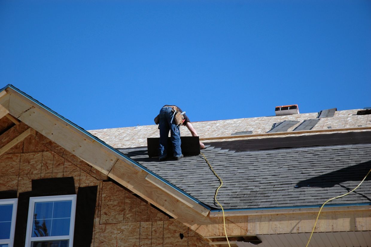 Worker installing shingles on a roof under clear sky.
