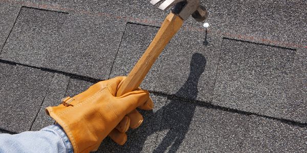 Person wearing glove holding hammer about to hit nail on shingled roof.