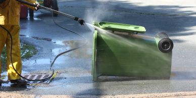 Person power washing a green trash bin outdoors.