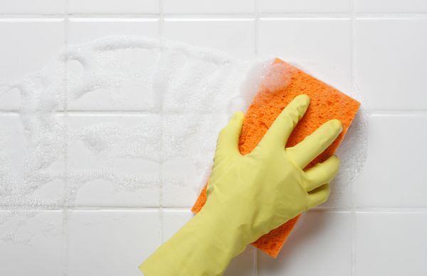 Hand in yellow glove cleaning white tiled surface with an orange sponge.