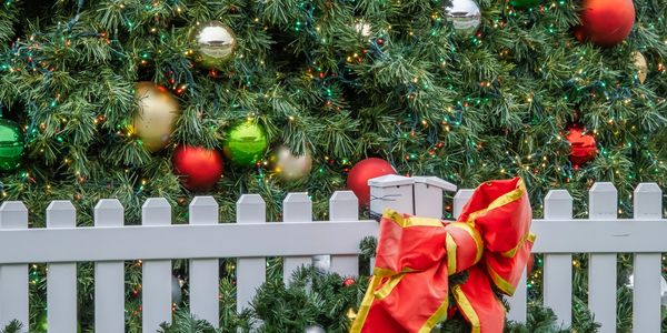 White fence decorated with Christmas garland and large red bow in front of a decorated Christmas tree.