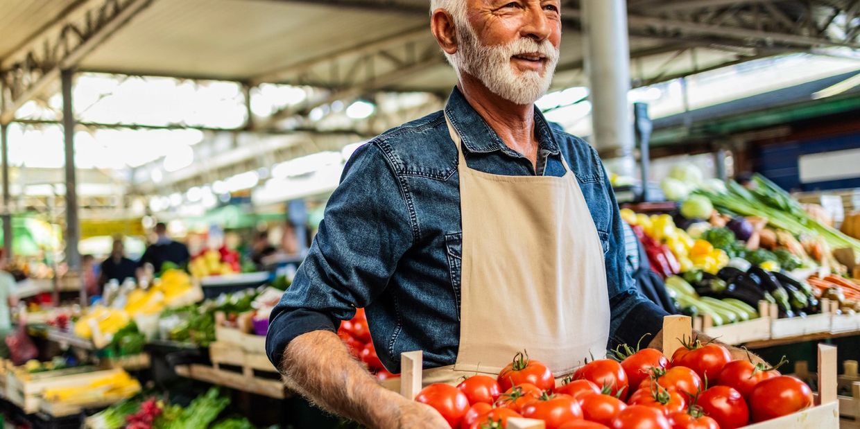 Smiling elderly man holding a crate of fresh tomatoes at a market.