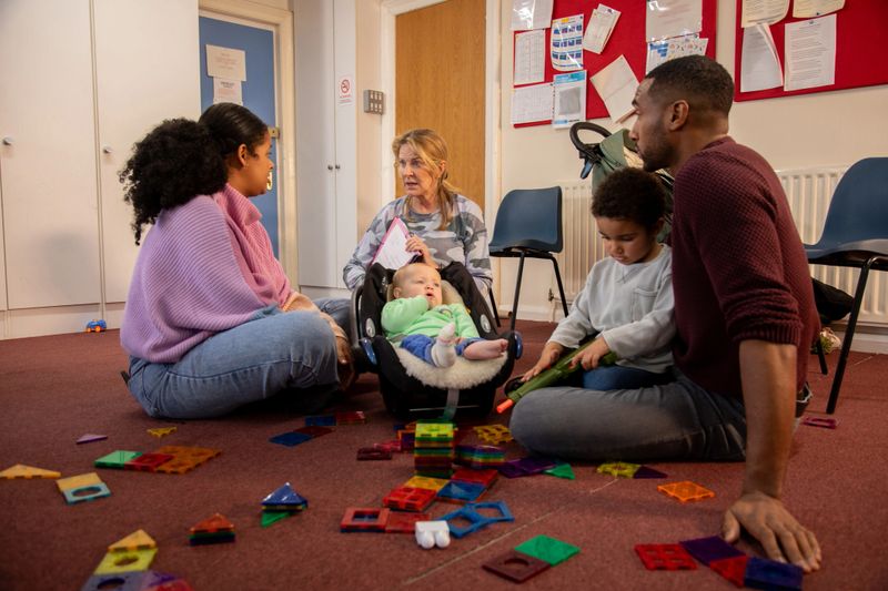 Full shot of a group of adults with their children having a meeting with a childcare worker. They are in a community centre surrounded by toys. The institute is located in Seghill, Northumberland.