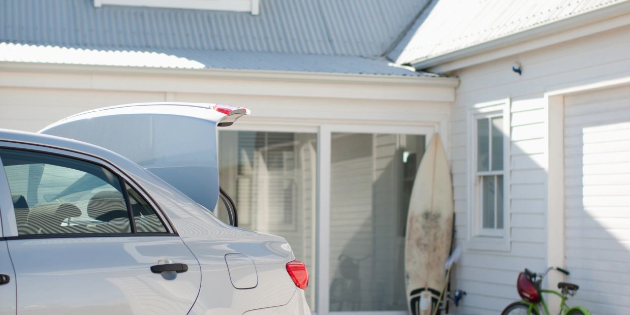 Car trunk open near a house with a surfboard and bicycle.