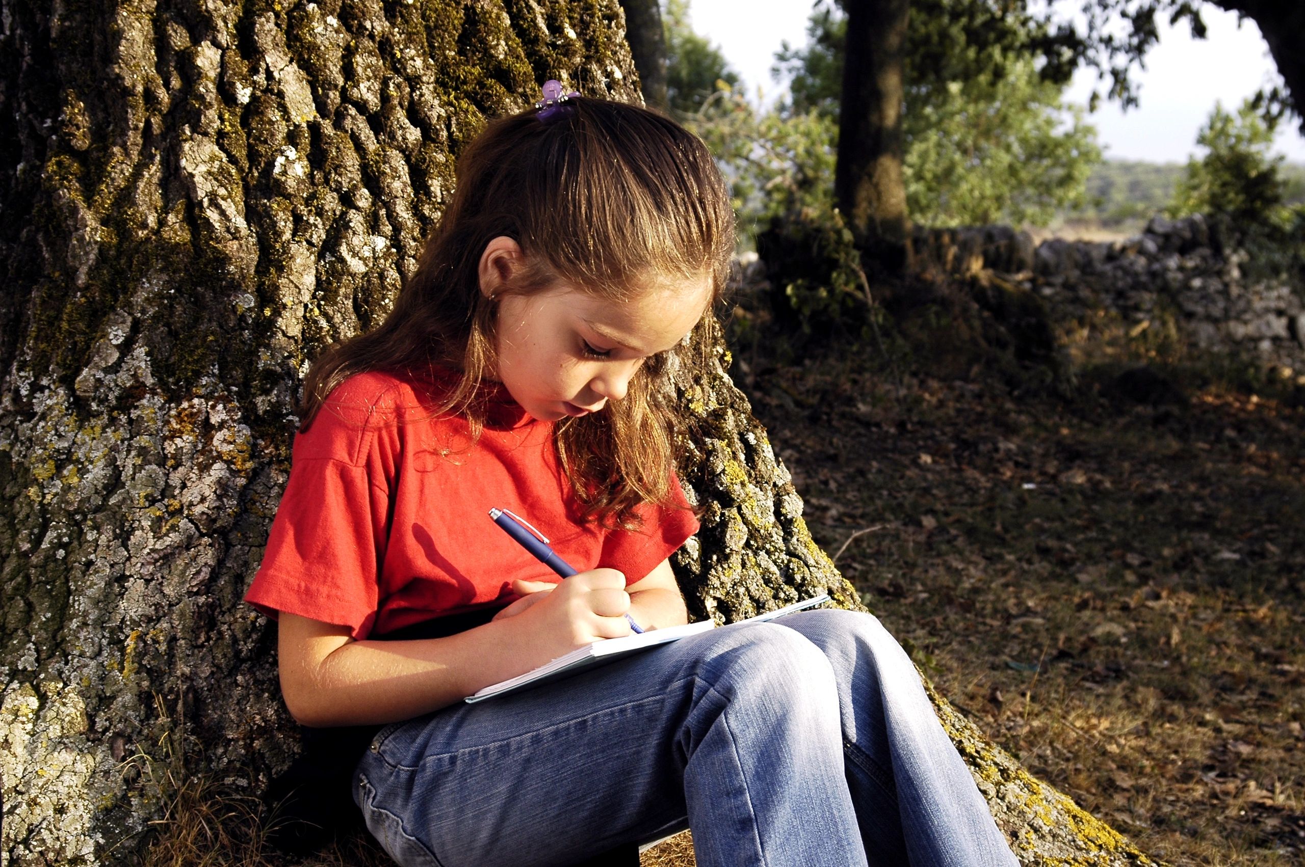 A young girl writing in a notebook while leaning against a tree.