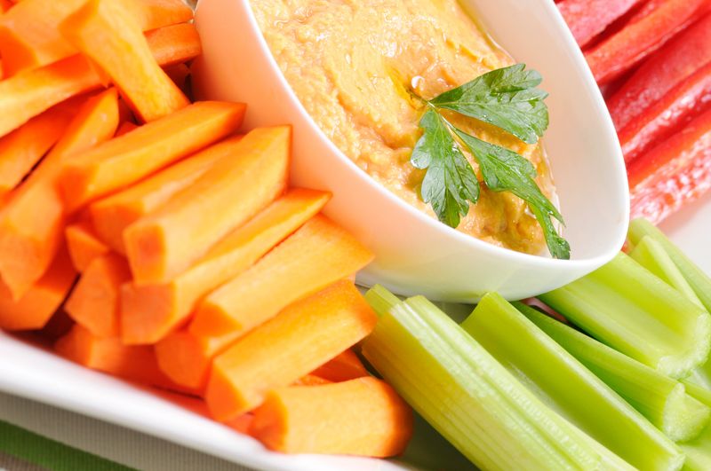 Macro close-up of a plate of fresh veggies with red pepper hummus for dip. The hummus is garnished with fresh Italian parsley.