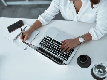 Person working on a laptop and taking notes at a clean white desk.