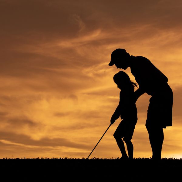Silhouette of adult teaching child golf at sunset.
