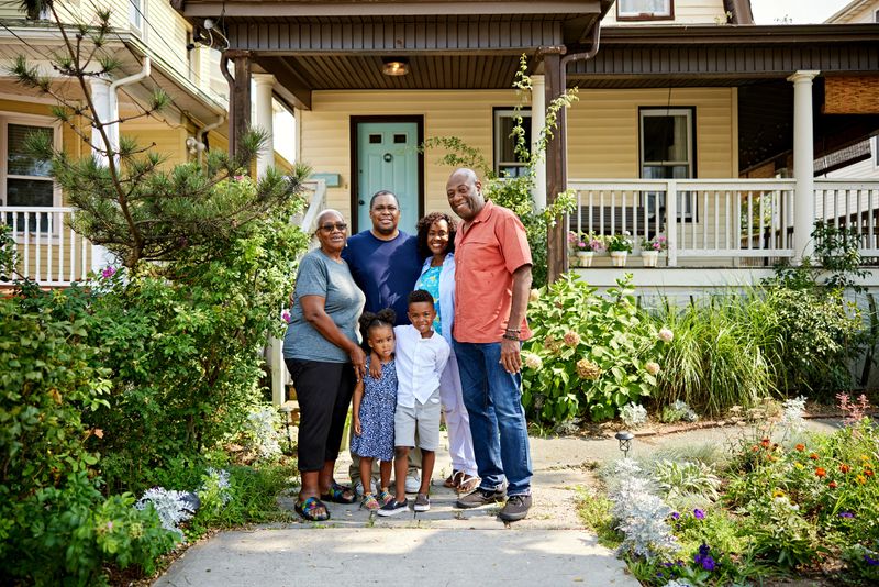 Full length view of young Black children, mature parents, and senior grandparents standing in garden of restored Victorian-style home and smiling at camera.