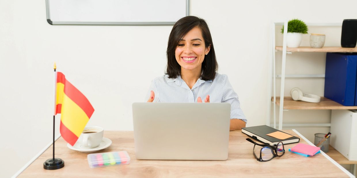 Smiling woman teaching Spanish online with a laptop and Spanish flag on desk.