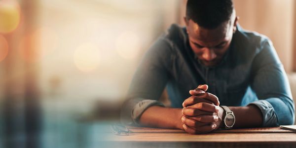 Man sitting with hands clasped, head bowed, in deep thought or prayer.