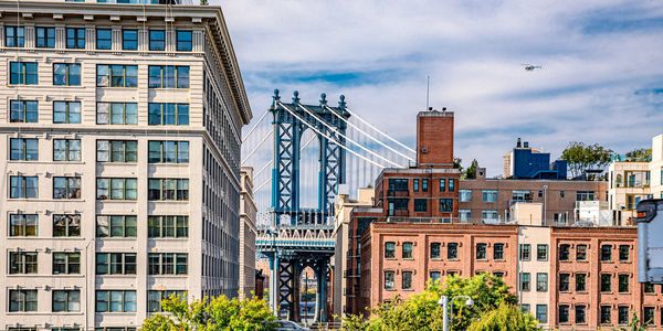 View of Manhattan Bridge framed by city buildings and greenery under a blue sky.