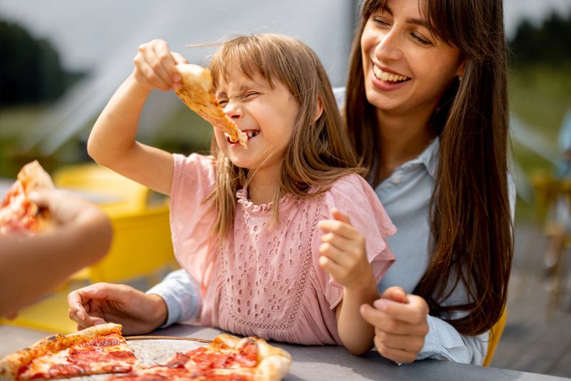 Cheerful mom with her little daughter eating pizza outdoors, visiting amusement park during a summer vacation
