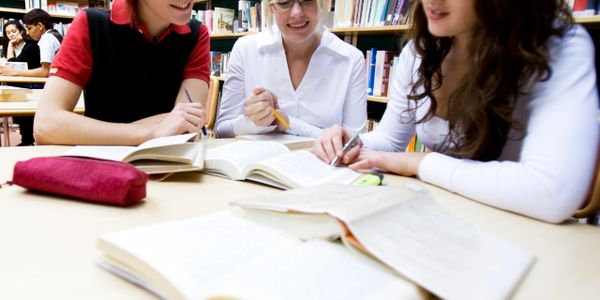 Three students studying together at a library table with books open.