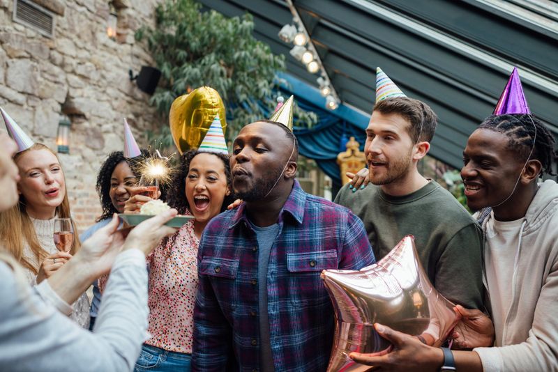 A group of friends wearing party hats, having a birthday party at a venue in Northumberland, North East England. One man is trying to blow out a sparkler on a cake that his friend is holding while his other friends watch in anticipation and celebrate.