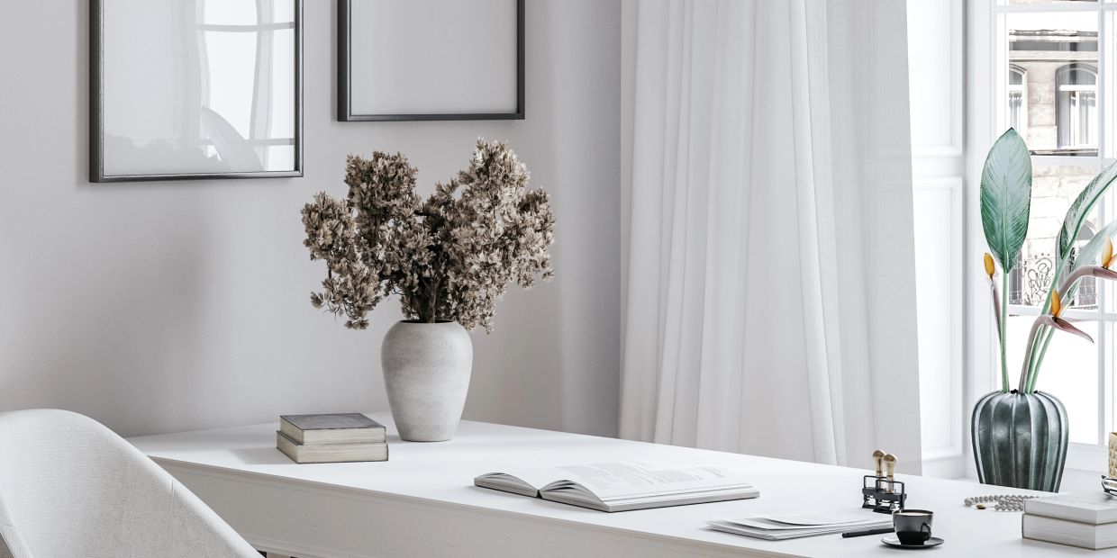 Minimalist white desk with dried flowers, books, and a coffee cup by a bright window.