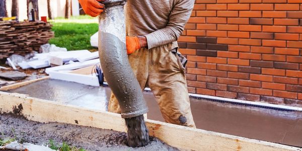 Worker pouring concrete into a wooden frame near a brick wall.
