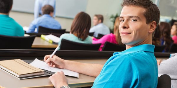 A young man in a blue shirt takes notes during a classroom lecture.