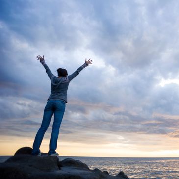 Person standing on rocks with arms raised under a dramatic cloudy sky at sunset.
