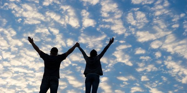 Two people holding hands with arms raised against a blue sky with scattered clouds.