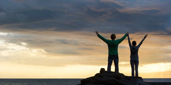 Two people standing on rocks by the ocean with arms raised at sunset.