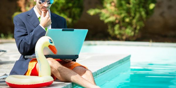 Man in suit jacket and swim trunks working on laptop by poolside.
