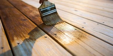 A hand applying golden stain to a wooden deck with a brush, captured in warm sunlight.