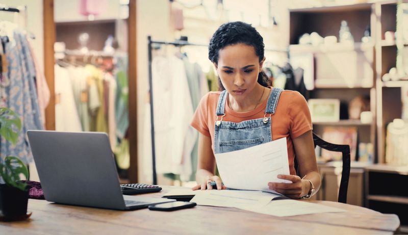 Business owner, clothes store and documents of startup debt, bills or financial report of e commerce sales on laptop. Entrepreneur, seller or confused woman reading paperwork, taxes or retail budget