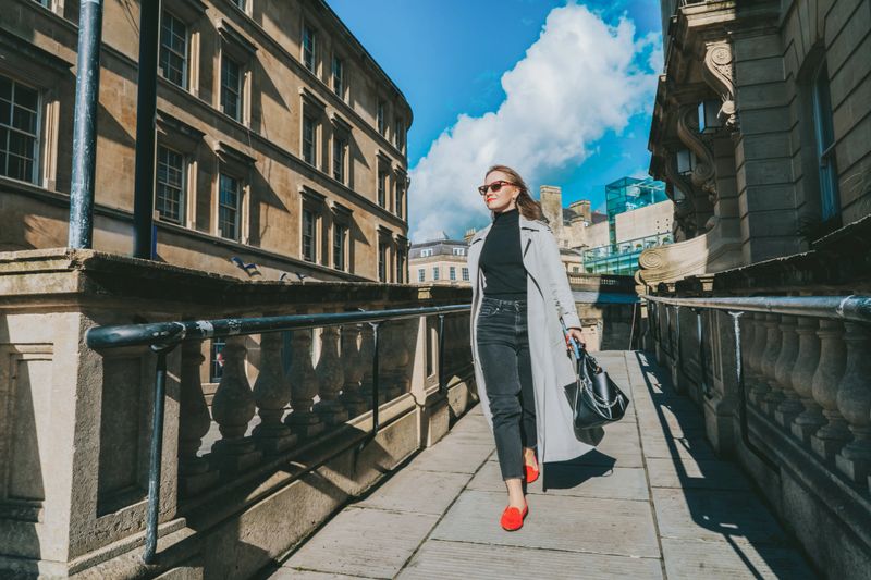 Outdoor autumn portrait of young elegant fashionable woman wearing trendy sunglasses, gray color coat, black jeans and turtleneck walking on street of European city in sunny day. street style fashion