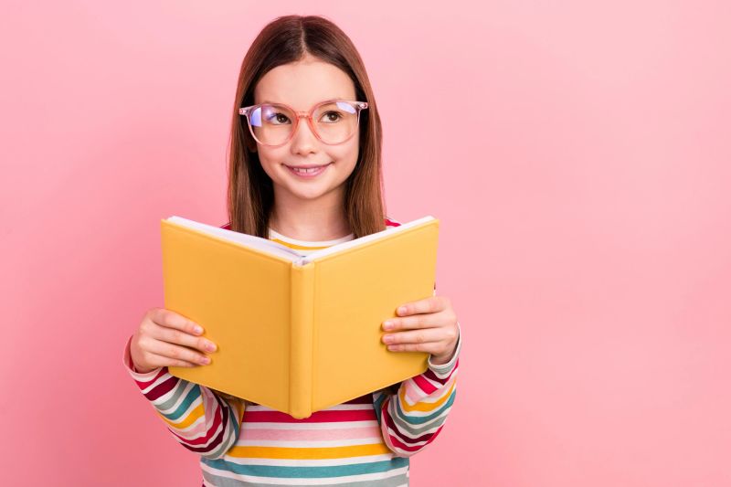 Photo of thoughtful little young schoolgirl kid reading favorite novel literature minded look empty space isolated on pink color background.
