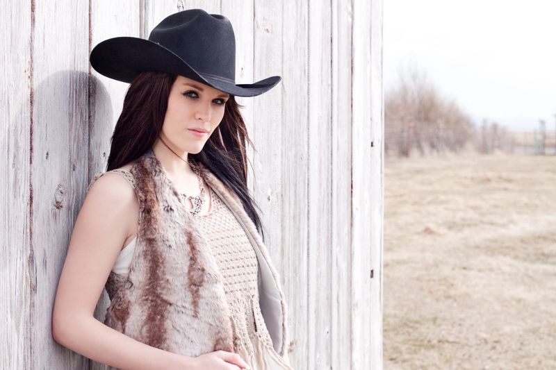 "Beautiful cowgirl outdoors wearing cowboy hat and holding a lariat, leaning against an old rustic wood fence."