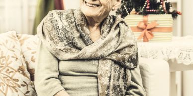Smiling elderly woman wrapped in a patterned shawl sitting indoors.