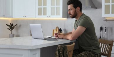 Man in camouflage pants working on a laptop in a kitchen.