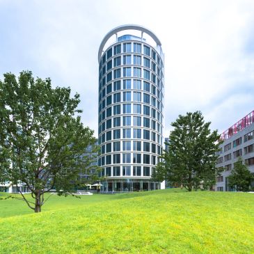 Modern cylindrical building surrounded by greenery under a cloudy sky.