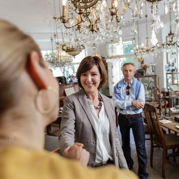Two women shaking hands in a vintage chandelier store with a man watching.