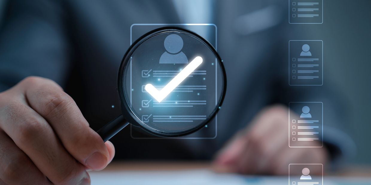 Person holding magnifying glass over a digital profile with a check mark.