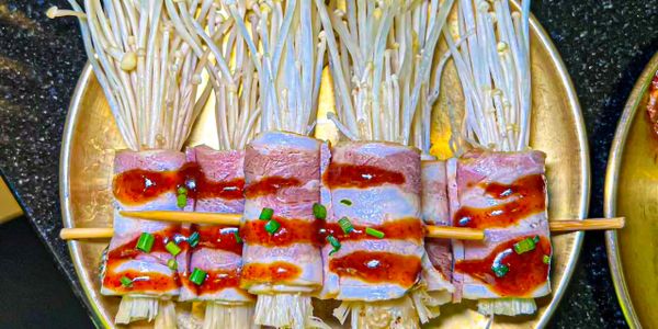 Grilled skewers with enoki mushrooms and bacon topped with sauce and chopped green onions.