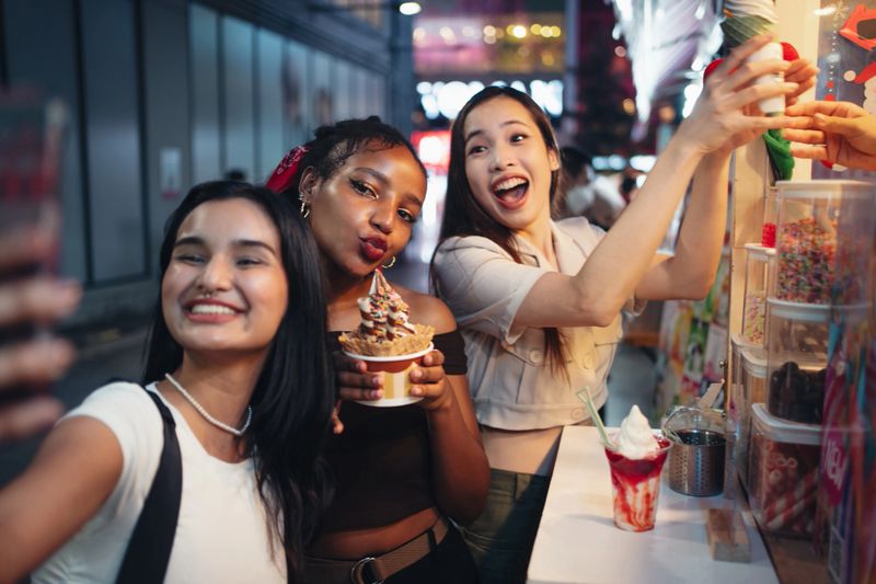 Joyful female friends are taking selfies with ice cream in the city at night.