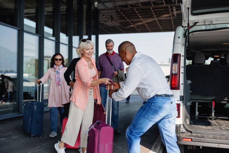 Full length side shot of a multicultural group of passengers waiting patiently for their suitcases that are being given by a Latin-American driver of the van that took them to the airport departure door on a sunny day.