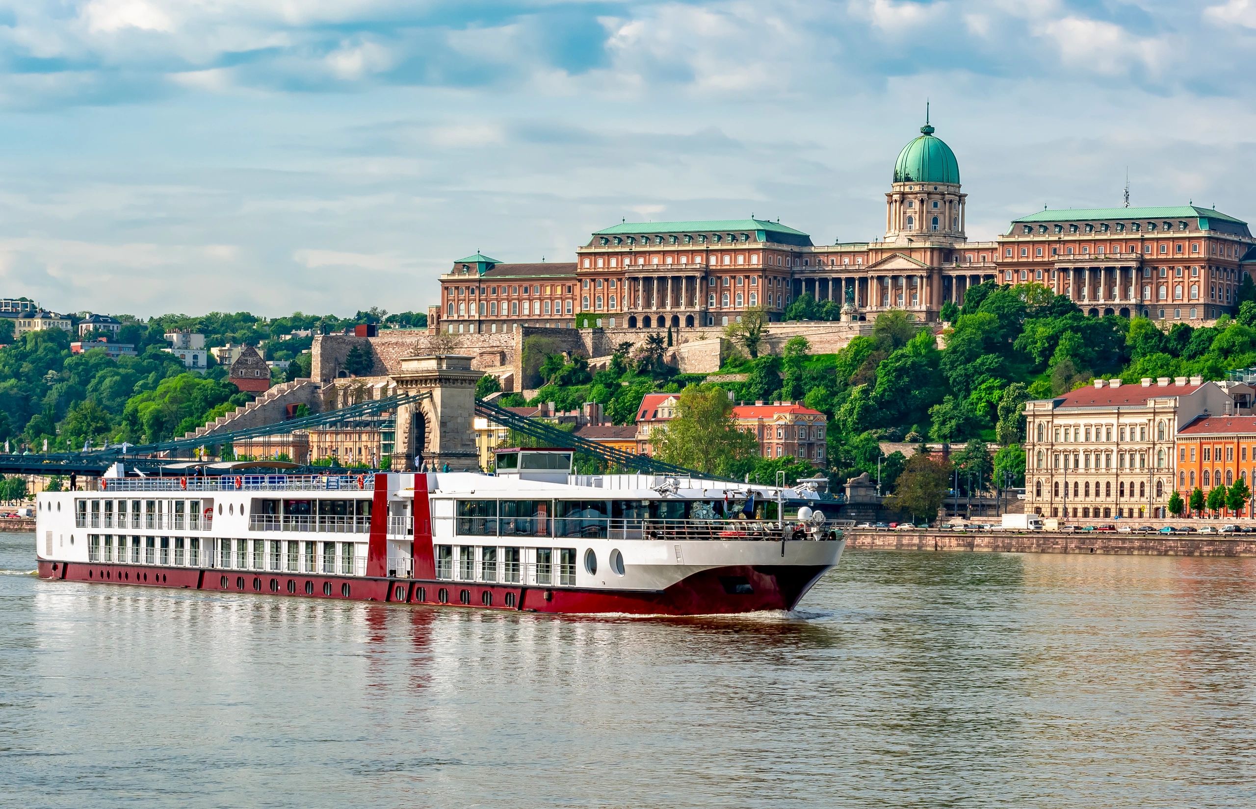 A large river cruise ship sailing near a historic castle with green domes.