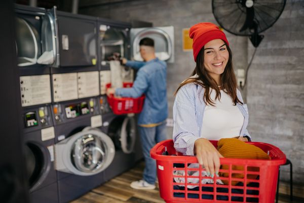 A woman holding a red laundry basket and smiling in a laundromat.