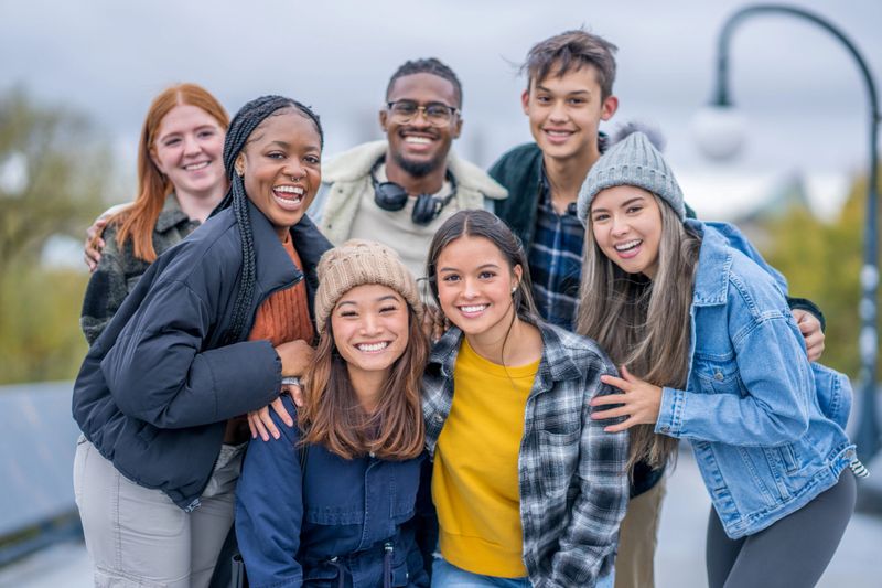 A large. group of University students huddle in closely together as they pose for a portrait.  They are each dressed casually in warm fall layers and are smiling.