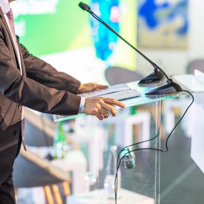 Person in suit giving a speech at a podium with microphones.