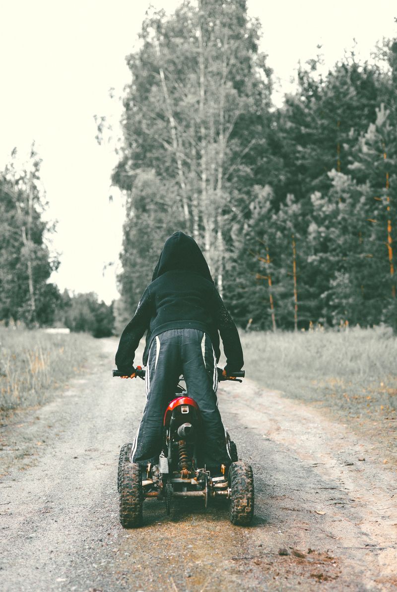 A child boy stands on an ATV wearing a black hoodie with his back to the camera. Quad bike. In the forest on a dirt road, a child stands riding his ATV. A child riding a small quad bike..