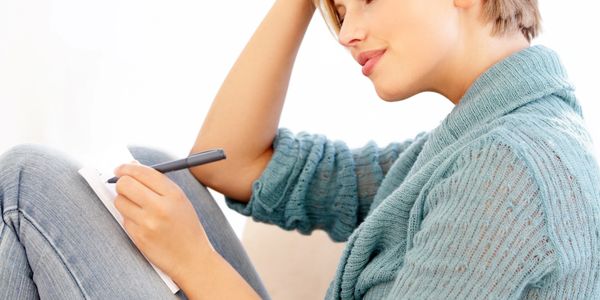 Woman in blue sweater writing in a notebook with a pen.