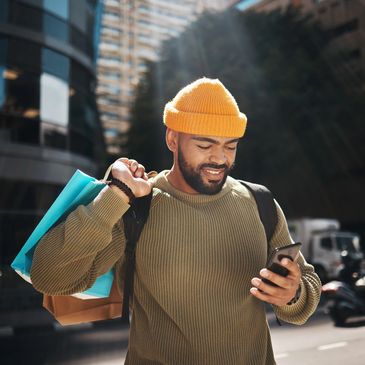 A man looking down at his phone while holding shopping bags over his shoulder