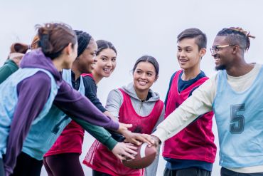 A diverse group of young people joining hands with a football on a field.
