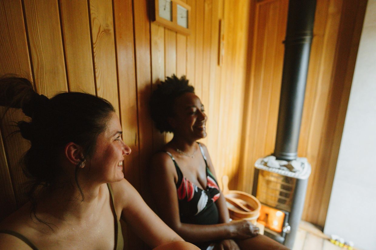 Two women smiling and relaxing inside a wooden sauna.