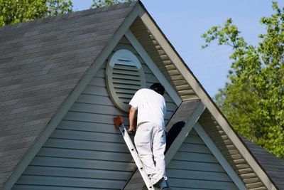 A man in white clothes climbs a ladder to the roof vent of a house.