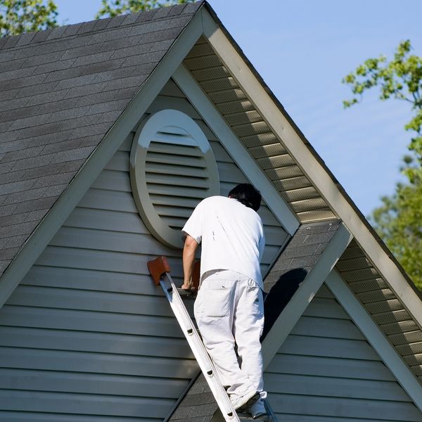 A man in white clothes climbs a ladder to the roof vent of a house.